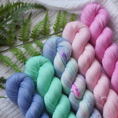 Five skeins of colorful yarn in blues, greens, and pinks on a textured surface with fern leaves.