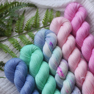 Five skeins of colorful yarn in blues, greens, and pinks on a textured surface with fern leaves.